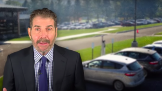 John Stossel stands in front of a row of cars | Stossel TV